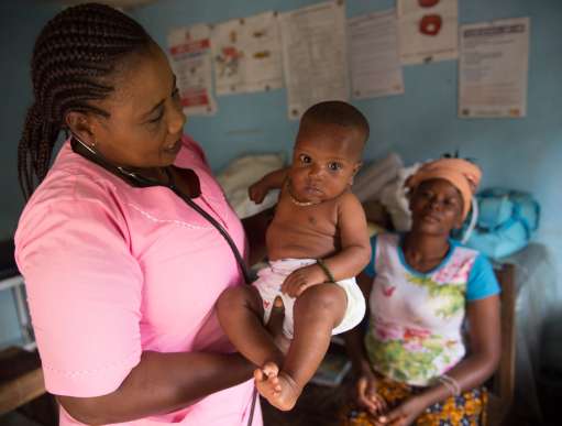 Nurse Judith stands holding a small baby, who is staring at the camera. The baby's mother sits, out of focus, in the background.