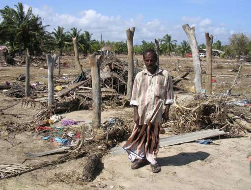 Man standing in beach strewn with clothing and remains of destroyed buildings